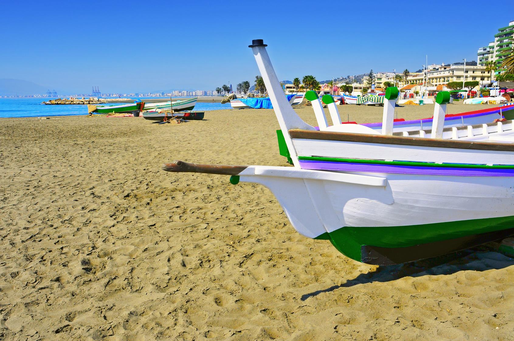 varias barcas sobre la arena de la playa, con los edificios de la costa y vista del puerto de Málaga al fondo