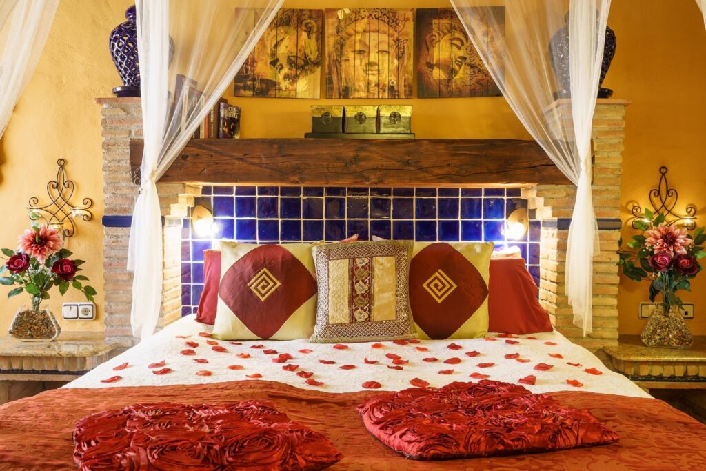 Cama de la habitación Orquídea Jacuzzi con imponente viga rústica de cabecero, cojines, alicatado... y pétalos de rosa sobre la cama, con colcha roja a juego y cuadros zen