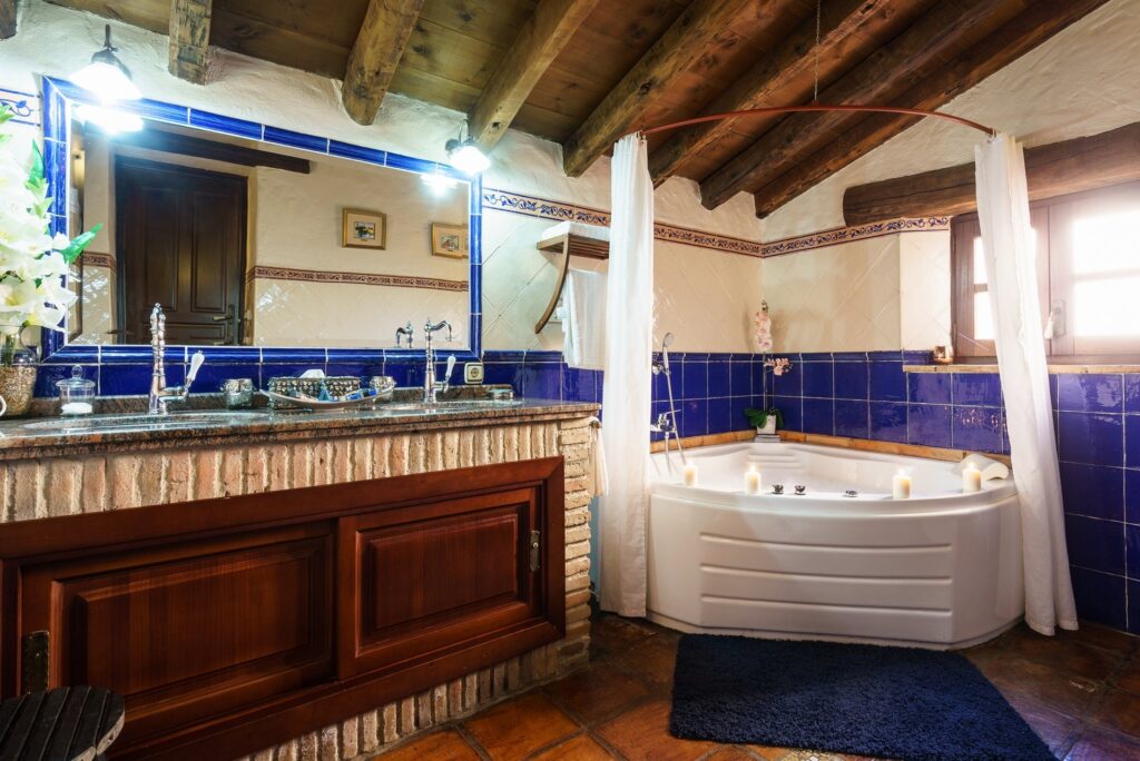Vista general del baño de la habitación Orquídea Jacuzzi, con doble lavabo, espectacular grifería rústica, espejo y resto de enseres típicos, con el jacuzzi al fondo.