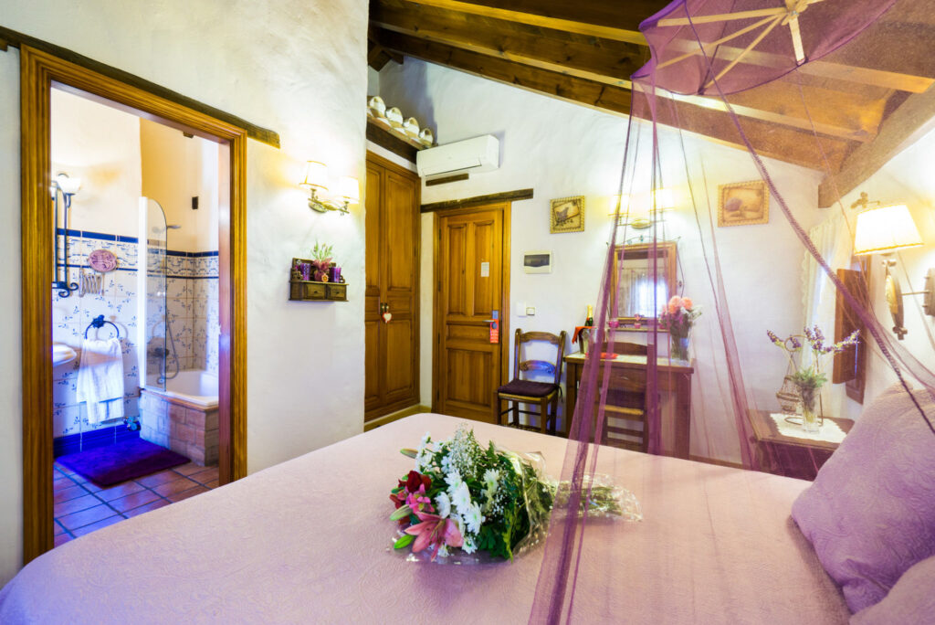 Cama con dosel y velo de la habitación Lavanda Romántica, con ramo de flores sobre la cama, acceso al baño en suite y mobiliario al fondo