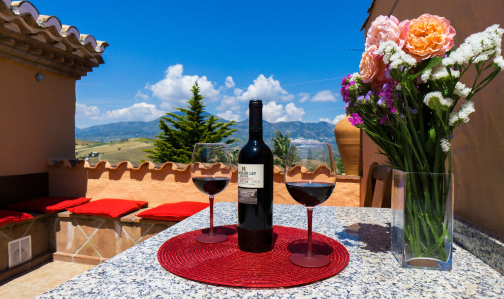 Botella de vino tinto con dos copas servidas, sobre bandeja roja con ramo de flores naturales, sobre mesa, en la terraza de la habitación MIMOSA JACUZZI