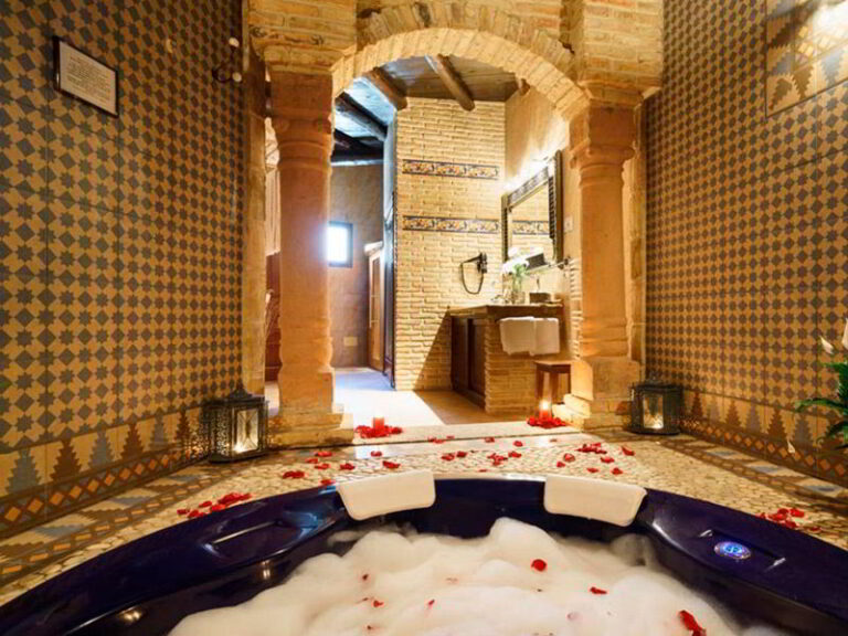 Jacuzzi romántico con espuma y pétalos de rosa dentro de la habitación Jazmín SPA Exclusive, al fondo acceso entre columnas desde la habitación