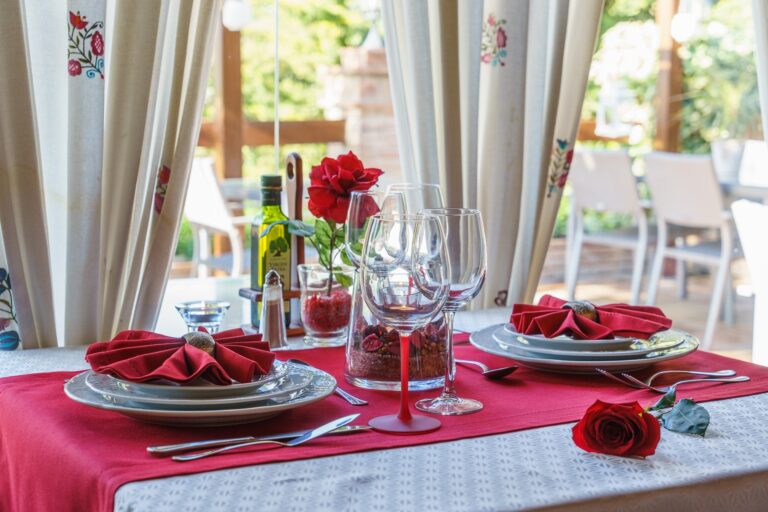 Mesa montada para dos comensales en restaurante acristalado, con detalles románticos: rosa roja sobre la mesa, etc