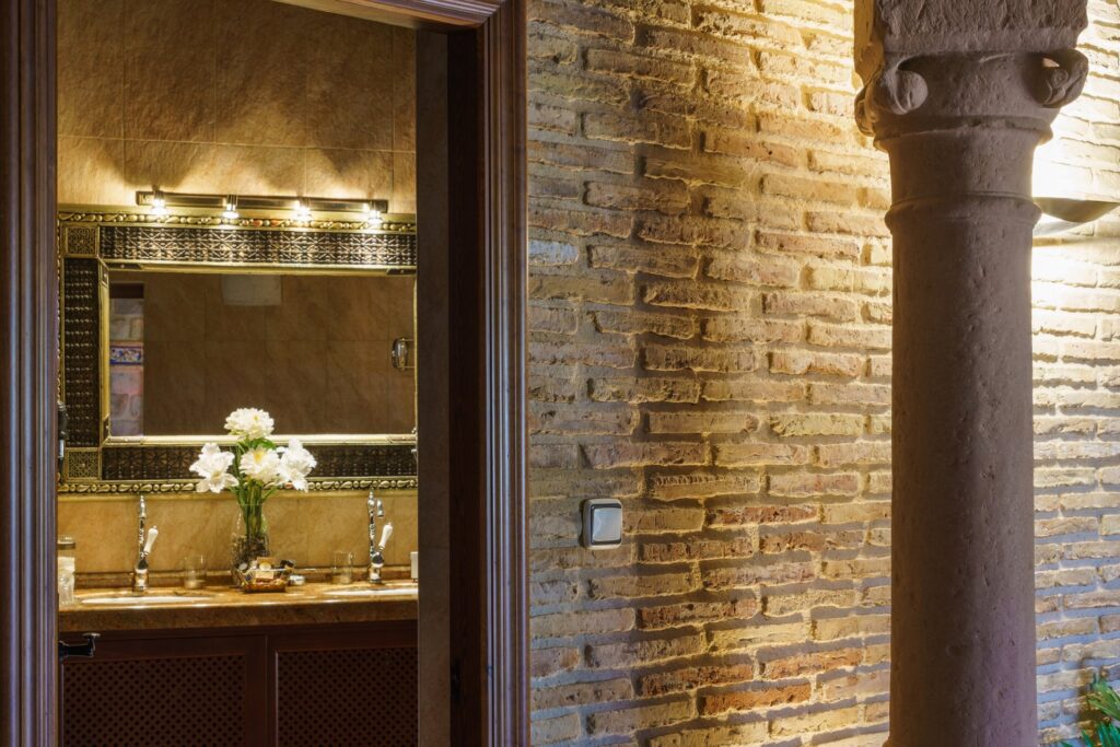 Entrada al baño de la habitación Jazmín SPA Exclusive con detalle de la pared estilo rústico, columna, y espejo con ramo de flores blancas al fondo