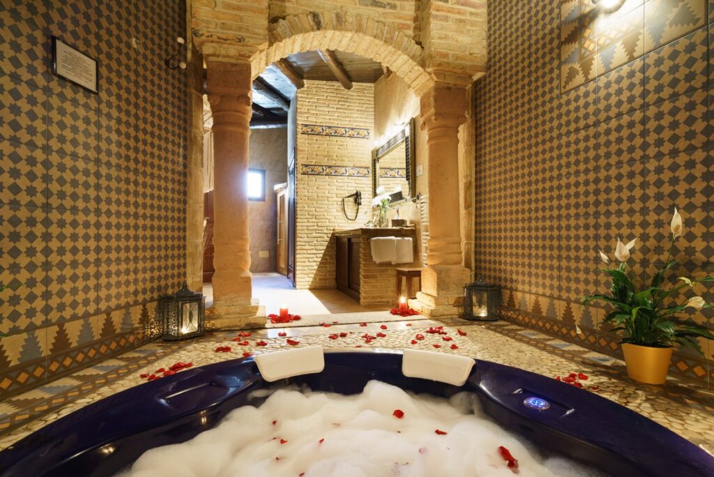 Jacuzzi romántico con espuma y pétalos de rosa dentro de la habitación Jazmín SPA Exclusive, al fondo acceso entre columnas desde la habitación