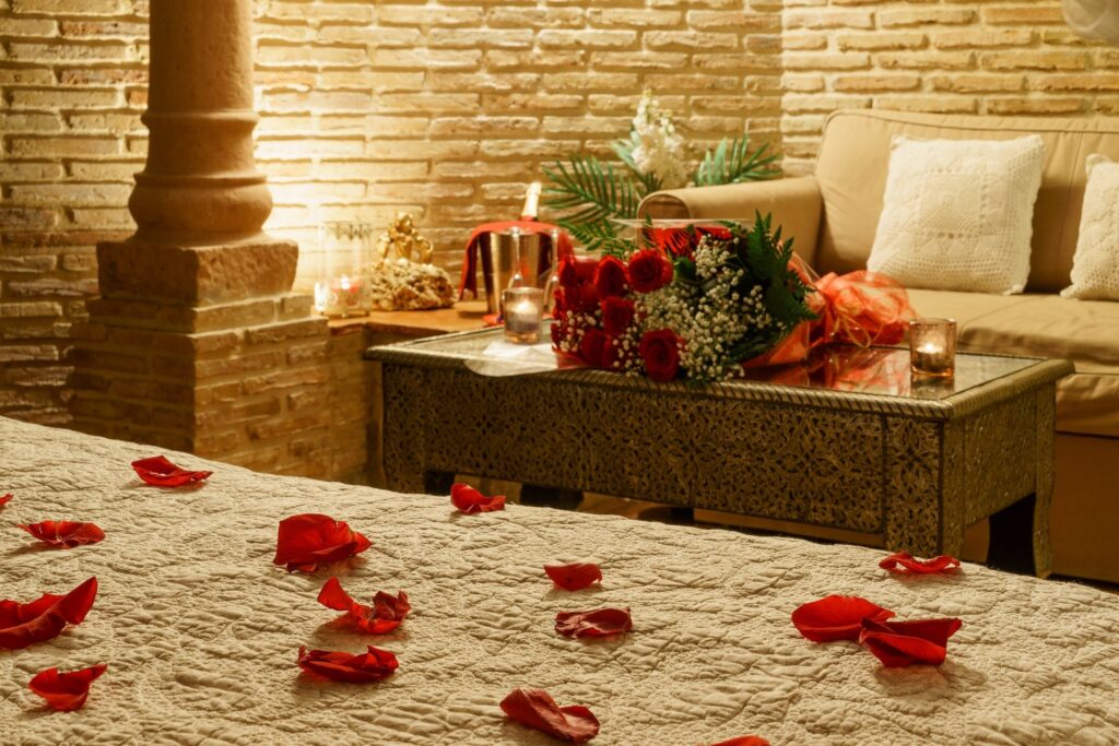 Detalle de mesa y sofá dentro de la habitación Jazmín SPA Exclusive, con velas y ramo de rosas encima, columna y pétalos de rosa esparcidos sobre la cama