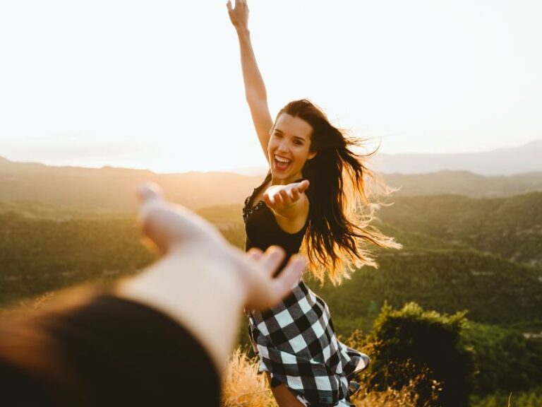 Chica joven feliz alzando los brazos en el campo, invitando a alguien de quien sólo se ve una mano